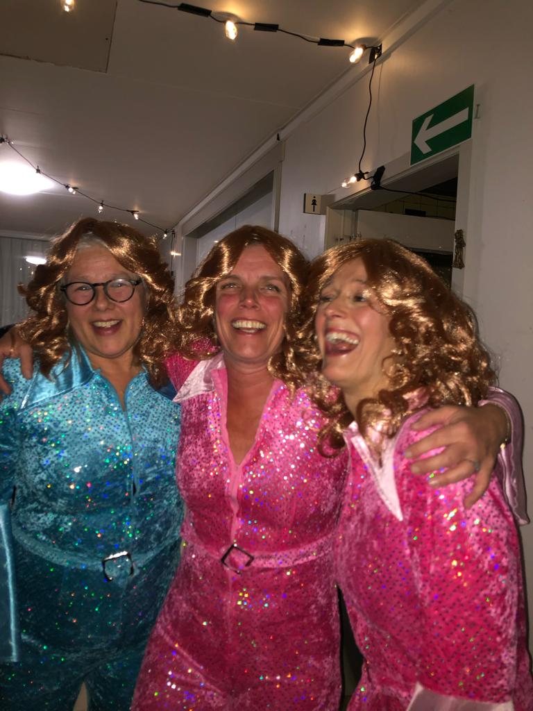 Three of the crew in sequin jumpsuits and curly wigs, arms around each other